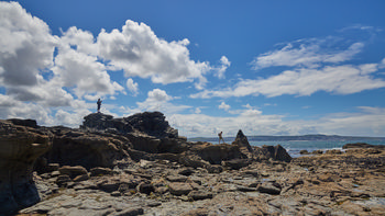 Godrevy rocks to St Ives This landscape photograph shows the rocky shoreline of Godrevy, looking toward St Ives in Cornwall, England, United Kingdom. Taken in the early afternoon during the summer season, the image captures the rugged rocks in the foreground extending out towards the sea, which is visible to the right, with gentle waves. Two people can be seen navigating the rocks, adding a sense of scale to the natural features. The sky is mostly clear with scattered clouds, and distant land is visible across the water, marking the coastal geography of Cornwall.